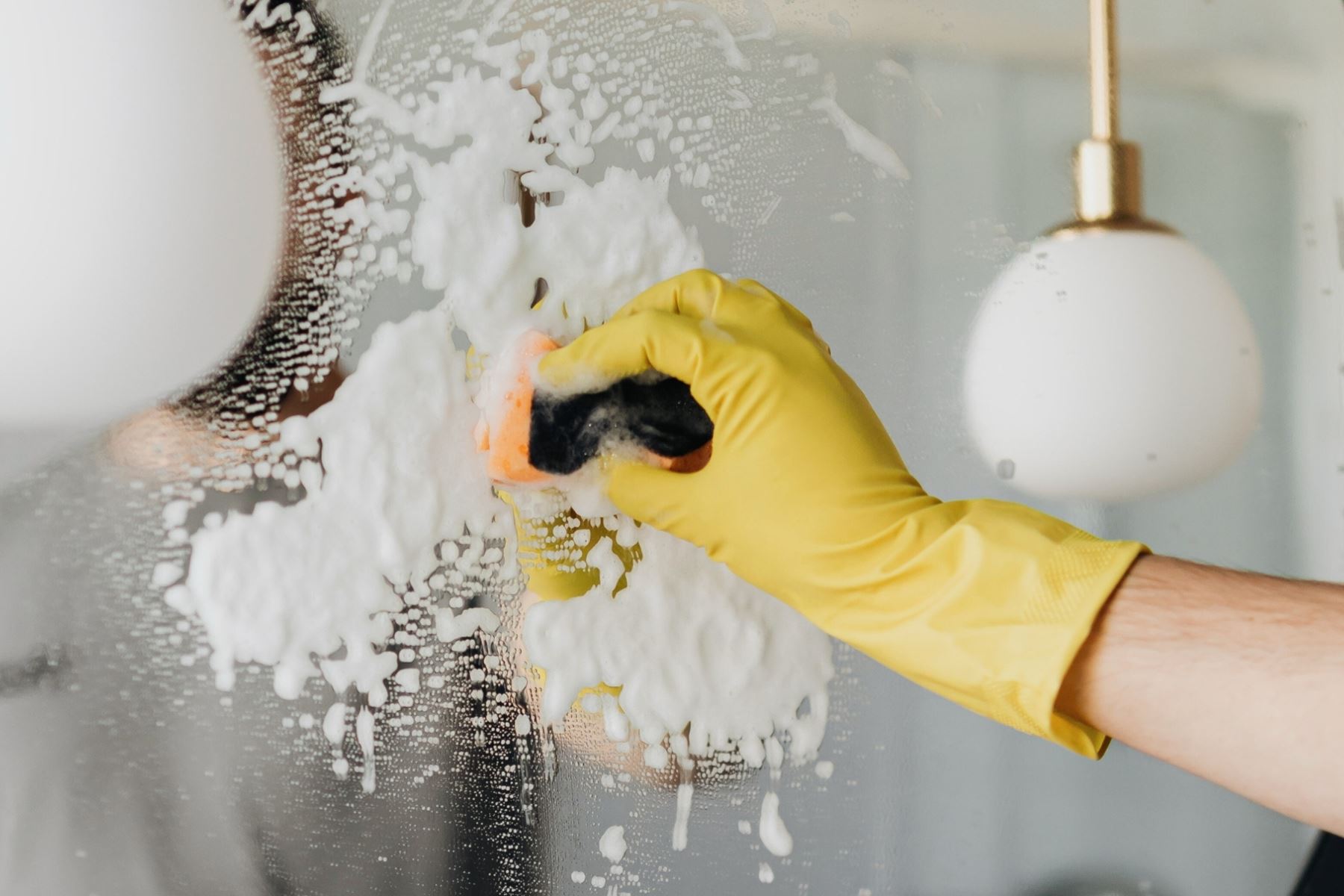 Hand i gul städhandske som putsar en spegel med tvättsvamp och löddrigt rengöringsmedel