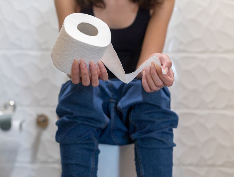 Kvinna som sitter på toaletten med nerdragna jeans och håller i en toalettrulle och drar ut en bit papper.