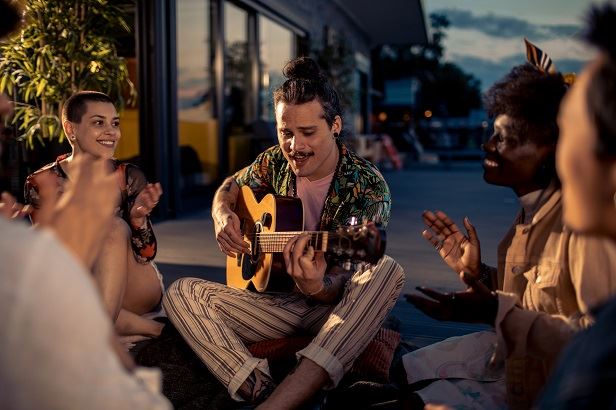 En man sitter på altangolvet utomhus och spelar gitarr för några vänner