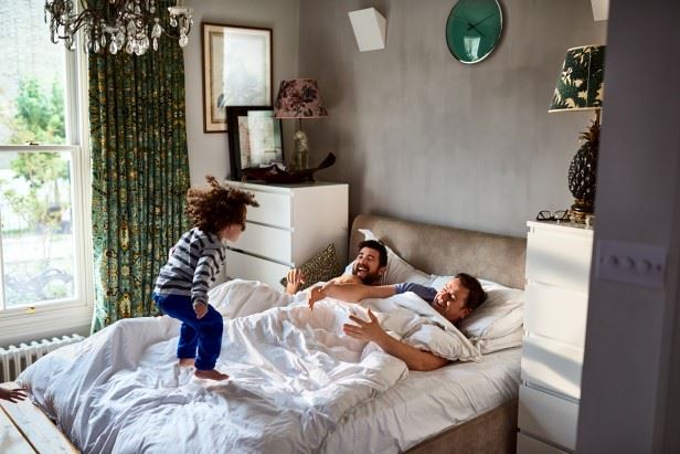 Två män ligger nerbäddade i sängen medan ett barn hoppar i sängen och skrattar